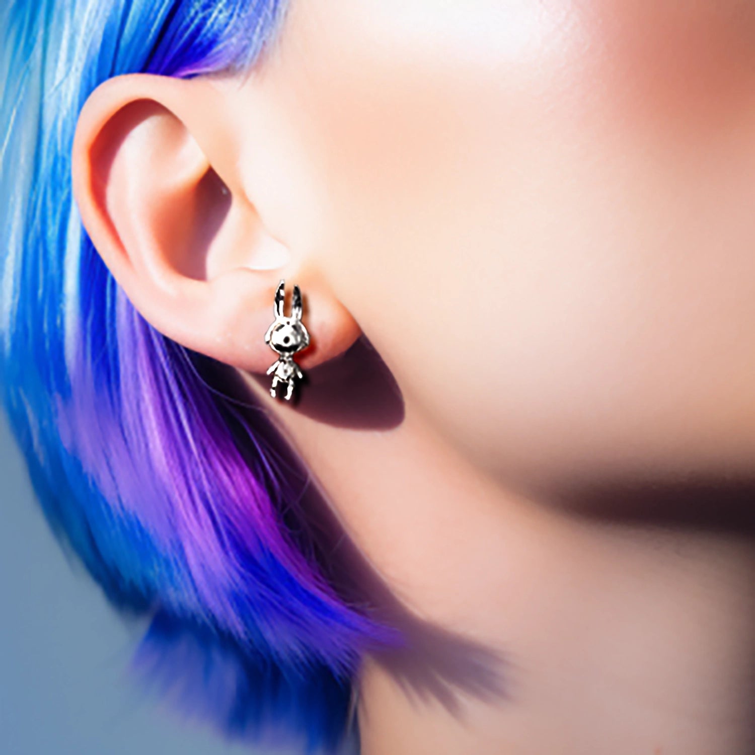 Close-up of an ear with a small astrobunny earring, hair with blue and purple tones.